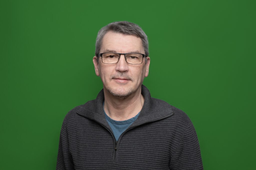Ein lächelnder Mann mit Brille in einem grauen Pullover vor einem grünem Hintergrund
