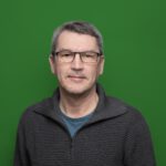 Ein lächelnder Mann mit Brille in einem grauen Pullover vor einem grünem Hintergrund