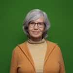 Eine lächelnde Frau mit einer Brille in einem orangem Oberteil vor einem grünem Hintergrund