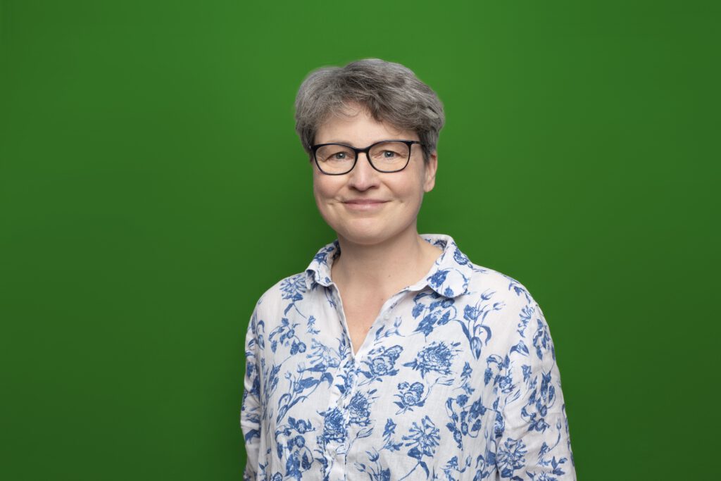 Foto von einer lächelnden Frau mit einer Brille in einem weißem Hemd mit blauen Blumen vor einem grünem Hintergrund