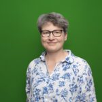 Foto von einer lächelnden Frau mit einer Brille in einem weißem Hemd mit blauen Blumen vor einem grünem Hintergrund