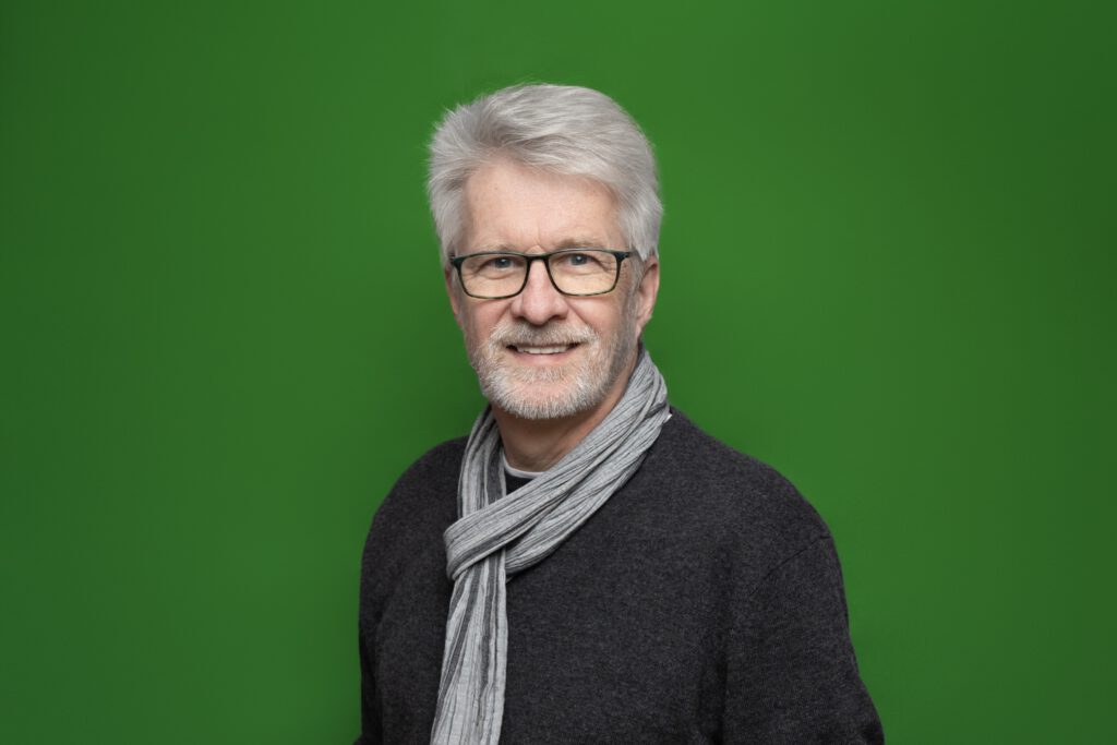Ein lächelnder Mann mit Brille in einem schwarzen Pullover mit einem grauen Schaal um den Hals vor einem grünem Hintergrund