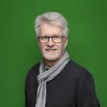 Ein lächelnder Mann mit Brille in einem schwarzen Pullover mit einem grauen Schaal um den Hals vor einem grünem Hintergrund