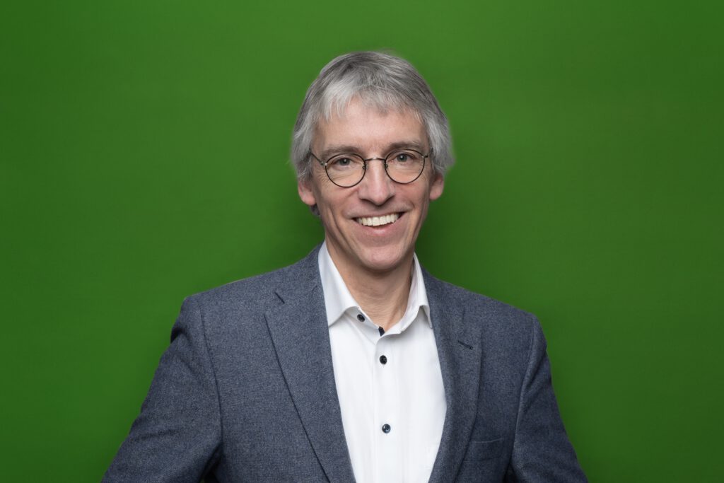 Ein lächelnder Mann mit Brille in einem grauen Anzug mit einem weißem Hemd vor einem grünem Hintergrund