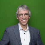 Ein lächelnder Mann mit Brille in einem grauen Anzug mit einem weißem Hemd vor einem grünem Hintergrund