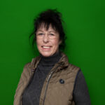 lächelnde Frau mit randloser Brille und Hochsteckfrisur mit grauem Rollkragenpullover und brauner Steppweste